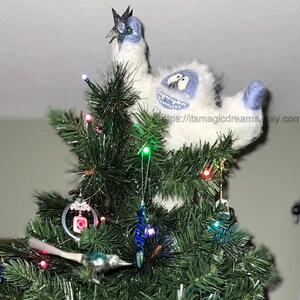 May include: A white and blue felt snowman ornament with a star on top, perched atop a green Christmas tree decorated with colorful lights and ornaments.
