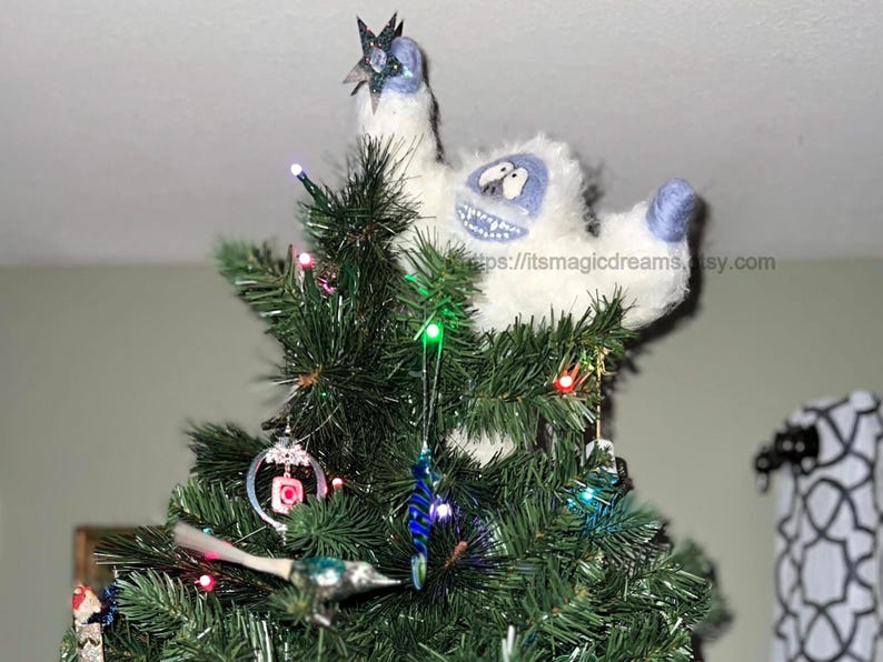 May include: A white and blue felt snowman ornament with a star on top, perched atop a green Christmas tree decorated with colorful lights and ornaments.
