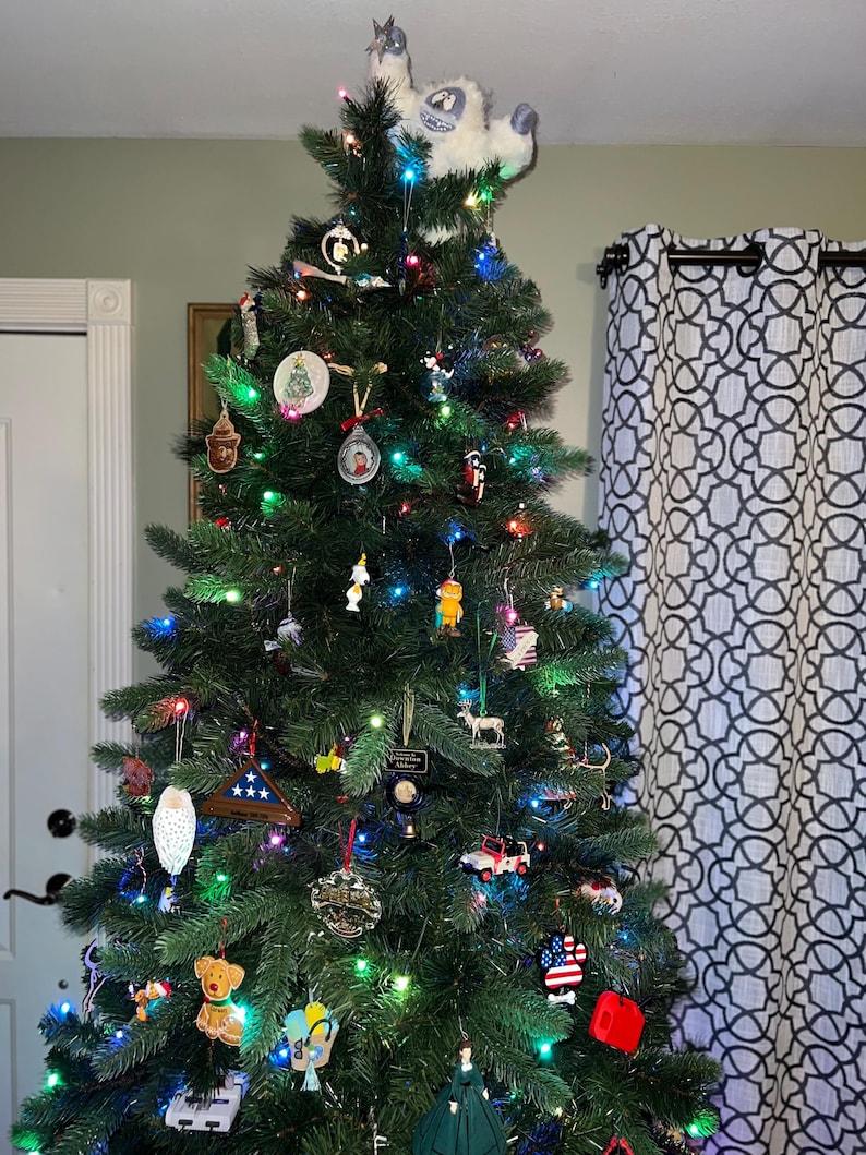 May include: A decorated Christmas tree with a snowman on top. The tree is decorated with ornaments, lights, and a variety of other decorations. The tree is in a room with a white door and a patterned curtain.