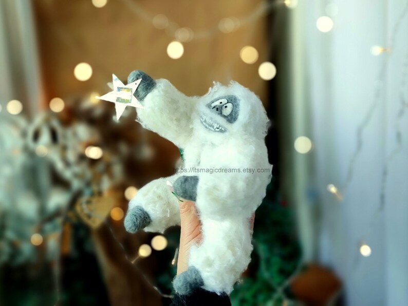 May include: A white plush yeti holding a silver star. The yeti has a gray face with black eyes and a black mouth. The yeti is sitting on a hand.