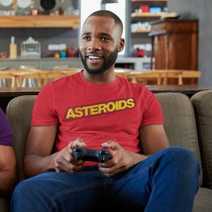 Puede incluir: Un hombre con una camiseta roja con la palabra "ASTEROIDS" impresa en amarillo en la parte delantera. Está sentado en un sofá y sostiene un mando de videojuego.