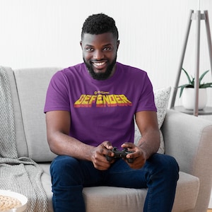 Puede incluir: Un hombre con una camiseta morada con el texto "Williams' Defender" en letras amarillas y rojas. Está sentado en un sofá y sostiene un mando de videojuego.