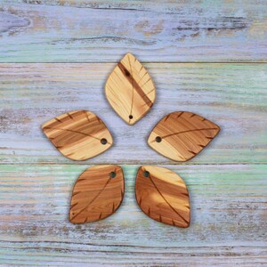 Può includere: Cinque ciondoli in legno a forma di foglia disposti a stella. Ogni pezzo presenta venature naturali del legno in marrone chiaro e scuro con una venatura centrale e linee laterali intagliate. Un piccolo foro circolare è praticato in alto.