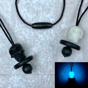 May include: Black and white skull-shaped pendants on black cords. The pendants have a pacifier-like design with a black ring and a faceted black bead. One skull glows blue in the dark. The pendants are displayed on a light gray surface.
