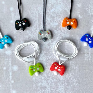 May include: Six colorful silicone game controller necklaces. The necklaces are on a white surface. The controllers are in the colors blue, green, red, orange, black, and gray.