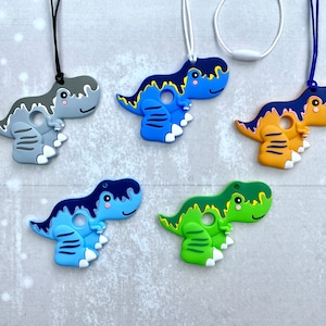 Puede incluir: Cinco collares de dentición de silicona con forma de dinosaurio en diferentes colores: gris, azul, amarillo, verde y azul claro. Cada collar presenta un dinosaurio con una superficie texturizada y un agujero en el medio.