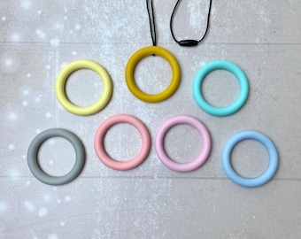 May include: A collection of colorful silicone teething rings in various pastel shades, including yellow, teal, pink, and blue. One mustard-colored ring is attached to a black cord. These rings are designed for babies.