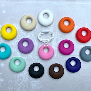 Puede incluir: Un conjunto de 15 anillos de dentición de silicona en varios colores, incluyendo amarillo, rojo, azul, verde, morado, rosa, marrón, negro, blanco y gris. Los anillos están dispuestos en un círculo sobre un fondo gris.