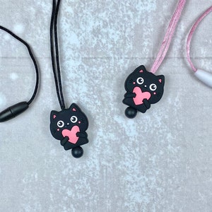 Può includere: Due collane con gatto nero e cuori rosa. Una collana ha un cordoncino nero e una chiusura nera. L'altra collana ha un cordoncino rosa e una chiusura bianca.