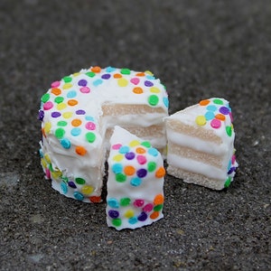 Puede incluir: Un pequeño pastel blanco con chispas de colores, cortado en tres trozos. El pastel está decorado con un glaseado blanco y chispas de colores.