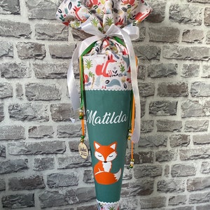 Könnte beinhalten: Eine grüne und weiße Schultüte mit einem Fuchsmotiv. Die Tüte trägt den Namen "Matilda" in weißer Schrift, eine Illustration eines schlafenden Fuchses und einen gemusterten Aufsatz mit Füchsen und Blättern. Sie ist mit Bändern und Perlen verziert.