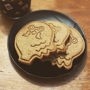 K&ouml;nnte beinhalten: Ein schwarzer Teller mit drei braunen Zuckerkeksen in Form von Geistern. Die Kekse haben ein wei&szlig;es Design. Der Teller steht auf einer Holzoberfl&auml;che neben einer Tasse Kaffee.