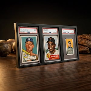 May include: A black display frame holding three vintage baseball cards featuring Mickey Mantle, Willie Mays, and Honus Wagner. The cards are in protective cases, with the players' images and card details visible. The frame sits on a wooden surface.