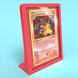 May include: A red plastic display stand with a clear plastic insert holding a holographic Pokemon trading card featuring Charizard, a fire-type Pokemon. The card has a red, yellow, and orange color scheme and features a picture of Charizard with its wings spread.