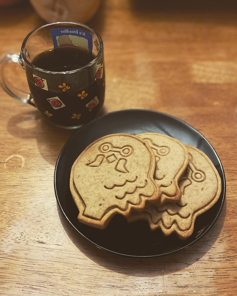 K&ouml;nnte beinhalten: Ein schwarzer Teller mit drei braunen Zuckerkeksen in Form von Geistern. Die Kekse haben ein wei&szlig;es Design. Der Teller steht auf einer Holzoberfl&auml;che neben einer Tasse Kaffee.