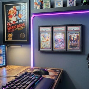 May include: A gaming setup with a wooden desk, keyboard, and mouse. A framed "Mario Bros." poster hangs on the wall, along with graded trading cards and a display case of video game cartridges. A headset and a Game Boy are also visible.