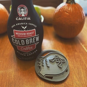 K&ouml;nnte beinhalten: Eine schwarze Flasche Califia Farms Cold Brew Coffee mit dem Text "100% Arabica Coffee" und "Medium Roast - Bold & Balanced - Cold Brew Coffee" auf dem Etikett. Die Flasche steht auf einer Holzoberfl&auml;che neben einem grauen 3D-gedruckten Objekt.