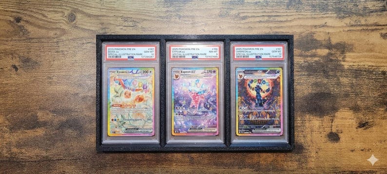 May include: A black framed display case holding three Pokemon trading cards. Each card is encased in a protective holder with a label at the top. The cards feature colorful illustrations and text.