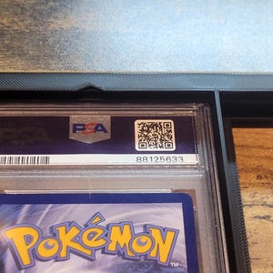 May include: A graded Pok&eacute;mon trading card in a protective case. The case has a PSA label with a QR code and serial number. The card is partially visible, showing the Pok&eacute;mon logo and a blue background.