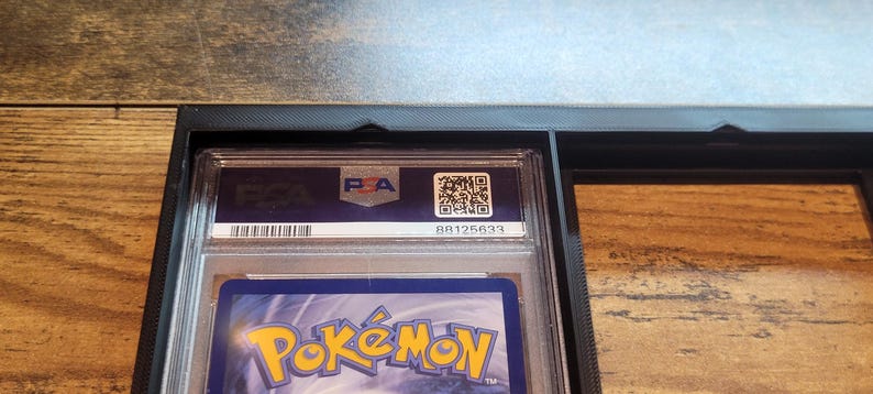 May include: A graded Pok&eacute;mon trading card in a protective case. The case has a PSA label with a QR code and serial number. The card is partially visible, showing the Pok&eacute;mon logo and a blue background.
