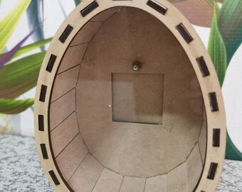 May include: A wooden egg-shaped box with a square opening and a small metal ball inside. The box is constructed from light brown wood with dark brown rectangular accents around the top edge. The interior is also light brown.