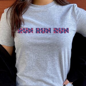 Könnte beinhalten: Graues T-Shirt mit dem Text "Run Run Run" in blauen Buchstaben mit einem roten und blauen Donut-Muster. Das Shirt wird von einer Person getragen.