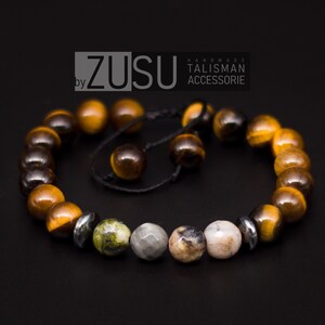 Peut inclure: Un bracelet en perles brunes et dorées avec un cordon noir et une perle argentée. Le bracelet a une perle de pierre verte et grise et une perle de pierre rose et grise. Le bracelet est fait à la main et porte le texte "ZUSU by TALISMAN ACCESSORIE" sur l'étiquette.