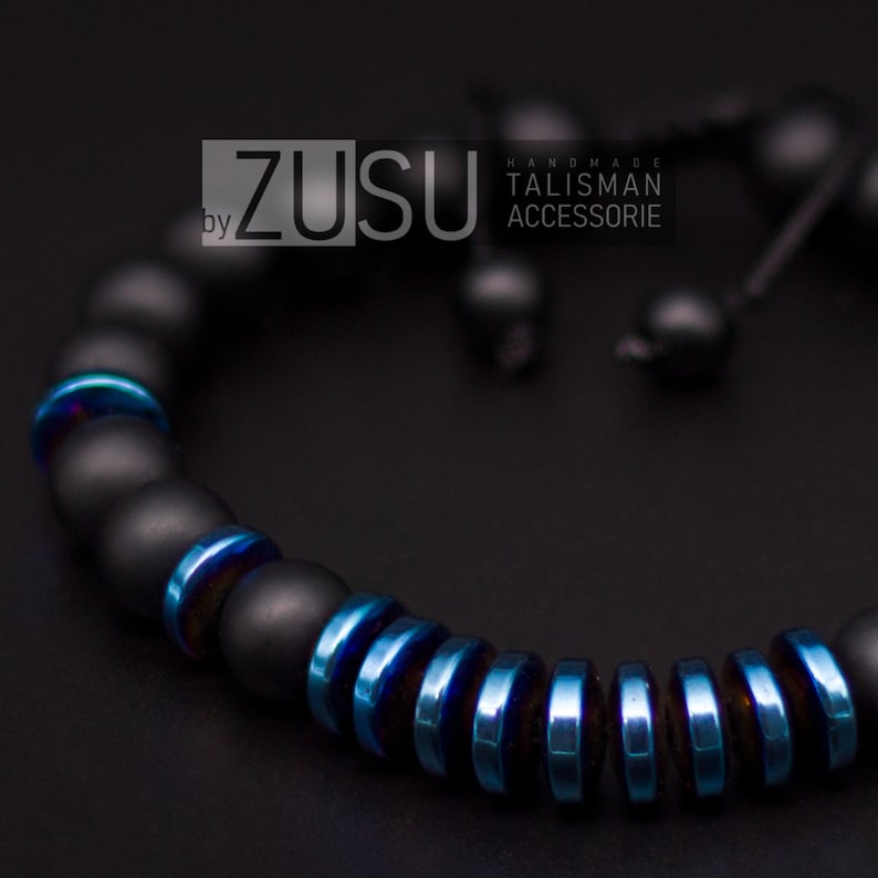May include: Black beaded bracelet with blue accents. The bracelet is made of matte black beads and blue metal beads. The bracelet is tied with a black cord. The text "by ZUSU HANDMADE TALISMAN ACCESSORIE" is visible in the image.