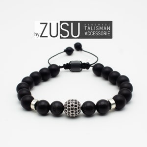May include: Black beaded bracelet with a silver charm featuring a black stone design. The bracelet has a black cord with a silver clasp. The bracelet is handmade and is labeled "ZUSU by HANDMADE TALISMAN ACCESSORIE".