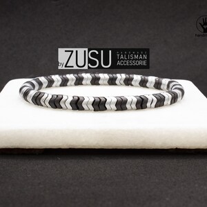 May include: Black and silver chevron patterned bracelet. The bracelet is made of small, flat, interlocking beads. The bracelet is on a white surface. The text "ZUSU by HANDMADE TALISMAN ACCESSORIE" is visible in the image.