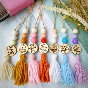 May include: Seven colorful wooden bead and tassel keychains with different inspirational messages. The messages include "Stay Safe", "Smile and Shine Bright", "Love with Joys Always", "Davis Family", "Dream Big Achieve More", "Stay Positive", and "I'm with You Always".