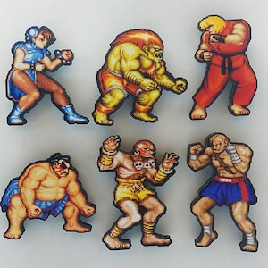 Puede incluir: Seis figuras de estilo pixel art de personajes de la serie de videojuegos Street Fighter. Las figuras están hechas de madera y están pintadas en colores brillantes. Los personajes incluyen a Ryu, Chun-Li, Sagat, Zangief, Blanka y E. Honda.