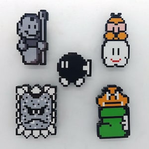 May include: Five pixel art character pins featuring a black and white character with a staff, a black and white bomb, a white character with a yellow character on top, a green and orange character, and a gray character with a black face.