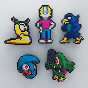 Puede incluir: Cinco pines de personajes de estilo pixel art. Una babosa amarilla, un pájaro azul, un monstruo verde, un personaje azul con una boca roja y una persona con un casco verde y blanco y una camisa rosa.