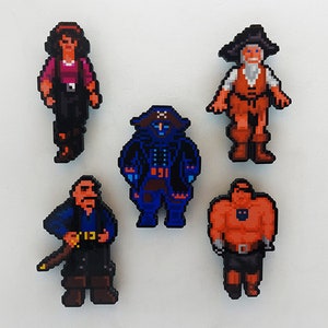 May include: Five pixel art style figurines of pirates. The figurines are made of felt and are approximately 7.5 cm tall. The pirates are wearing various outfits, including a red and white striped shirt, a blue coat, and a black and red dress.
