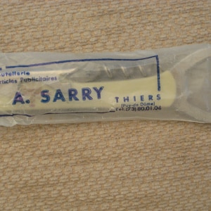 Pode incluir: Um pacote branco e azul contendo um abridor de garrafas prateado com saca-rolhas. O pacote tem o texto "Usines de Coutellerie et d'Articles Publicitaires A. SARRY THIERS (Puy-de-Dôme) Tel. (23) 80.01.04" impresso nele.