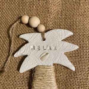 May include: White ceramic palm tree ornament with the word "Relax" and a seashell on the front. The ornament is hanging from a twine string with wooden beads.