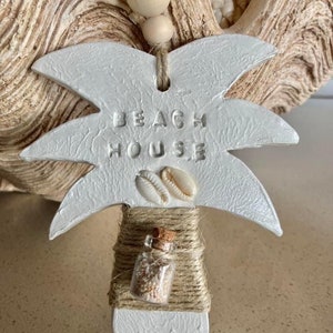 May include: White ceramic palm tree ornament with the words "Beach House" embossed on the trunk. The ornament has a small glass bottle filled with sand and two seashells attached to the trunk.