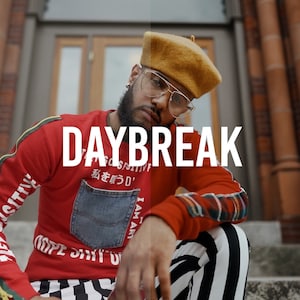 May include: A man wearing a red sweater with a blue denim pocket and black and white striped pants. The sweater has the text "DAYBREAK" and Japanese characters on it. He is wearing a brown beret and glasses.