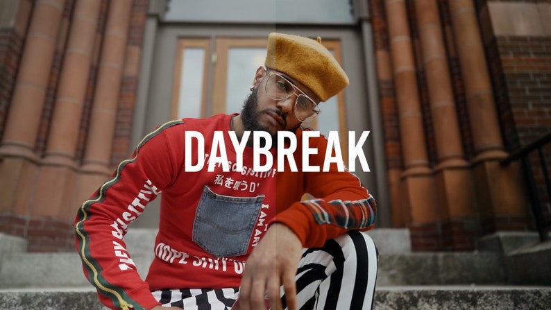 May include: A man wearing a red sweater with a blue denim pocket and black and white striped pants. The sweater has the text "DAYBREAK" and Japanese characters on it. He is wearing a brown beret and glasses.