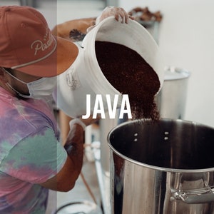 May include: A person wearing a tie-dye shirt and a red baseball cap with the word "Pacific" on it pours coffee beans into a large stainless steel container. The container is labeled "JAVA".