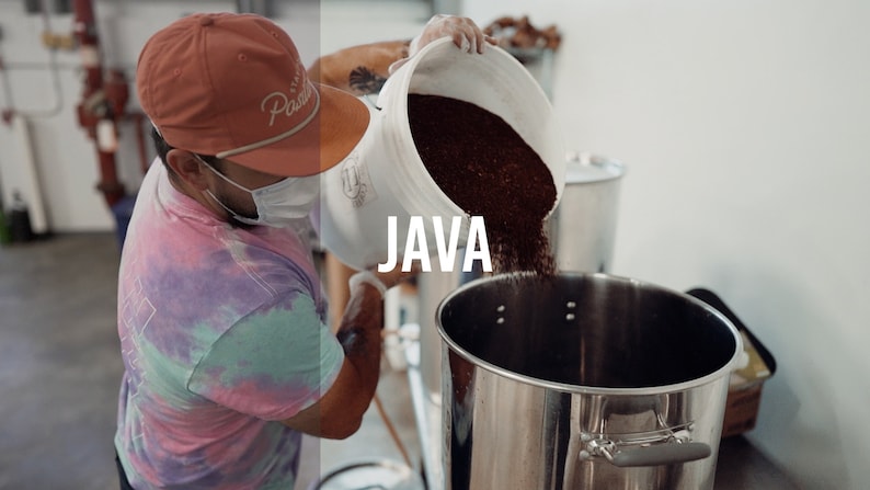 May include: A person wearing a tie-dye shirt and a red baseball cap with the word "Pacific" on it pours coffee beans into a large stainless steel container. The container is labeled "JAVA".