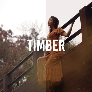 May include: A woman in a long, orange floral dress stands on a wooden bridge, holding onto the railing. The word "TIMBER" is printed in white letters on the bridge.