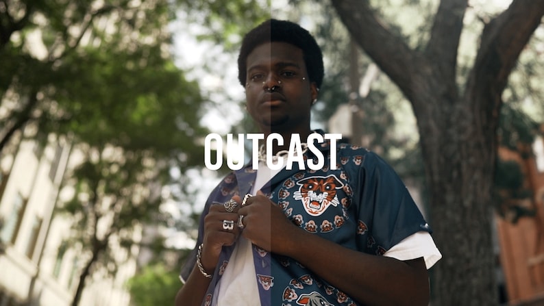 May include: A person wearing a blue and white patterned shirt with a tiger graphic. The shirt has the word "OUTCAST" printed on it.