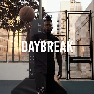 May include: A man wearing a black shirt with the word "DAYBREAK" printed on it, is holding a basketball in his right hand. He is standing on a basketball court with a chain-link fence in the background.