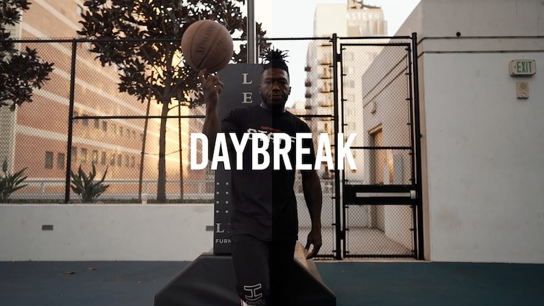 May include: A man wearing a black shirt with the word "DAYBREAK" printed on it, is holding a basketball in his right hand. He is standing on a basketball court with a chain-link fence in the background.