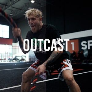 May include: A man wearing a black t-shirt with the word "OUTCAST" printed on it, black and red shorts, and black wristbands, is working out with a battle rope in a gym. The gym has a black wall with the words "1 SPORTS" printed on it.