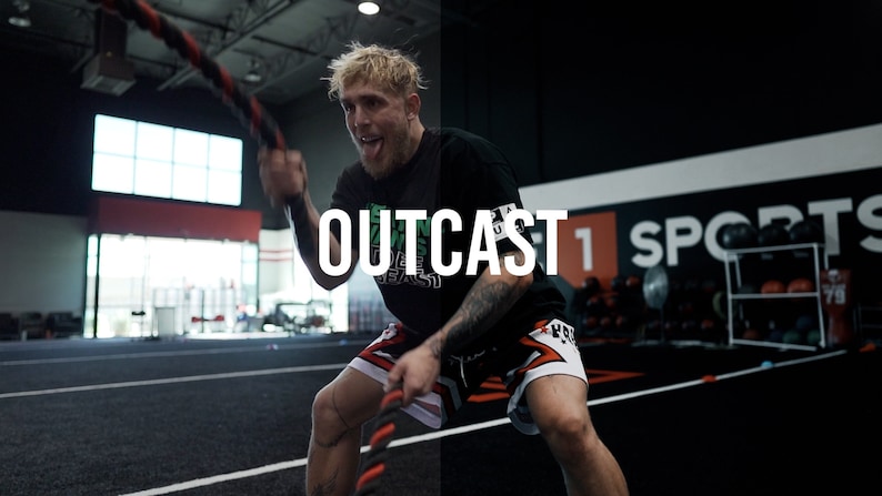 May include: A man wearing a black t-shirt with the word "OUTCAST" printed on it, black and red shorts, and black wristbands, is working out with a battle rope in a gym. The gym has a black wall with the words "1 SPORTS" printed on it.