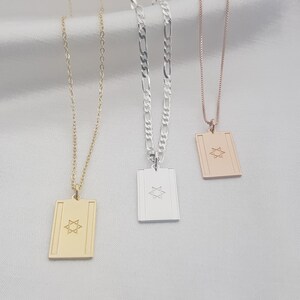 Könnte beinhalten: Drei Anhängerhalsketten in Gold, Silber und Roségold. Jeder rechteckige Anhänger zeigt ein Davidstern-Design. Die Halsketten hängen auf einem weißen, seidigen Hintergrund.