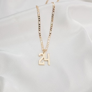 May include: A gold-coloured necklace with a chain and a pendant featuring the number 24. The necklace is displayed on a white, silky fabric. The chain has a textured design.